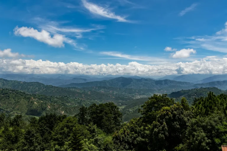 Panoramic view of lush green rolling hills and valleys in Dhulikhel, Nepal, under a bright blue sky with wispy clouds.