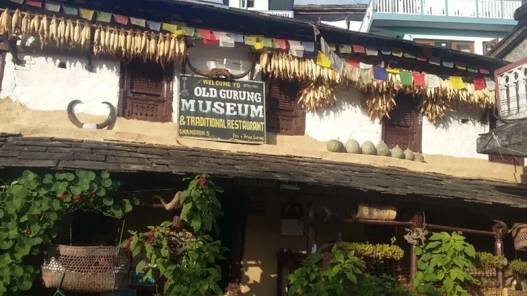 Traditional stone building in Ghandruk, Nepal, featuring a sign for the Old Gurung Museum, hanging rows of dried corn, and colorful prayer flags.