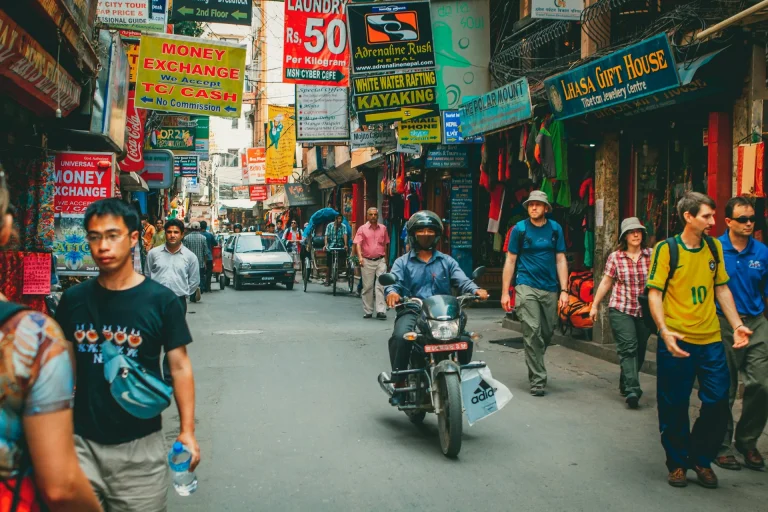 A crowded street in Thamel, Kathmandu, filled with pedestrians, a motorcyclist, and colorful signs for shops and money exchanges.