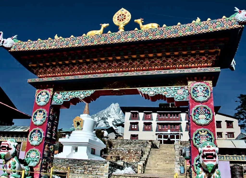 An ornate Tibetan Buddhist gateway with a golden Dharma wheel on top framing a snow-capped Himalayan peak and a white stupa in front of Tengboche Monastery.