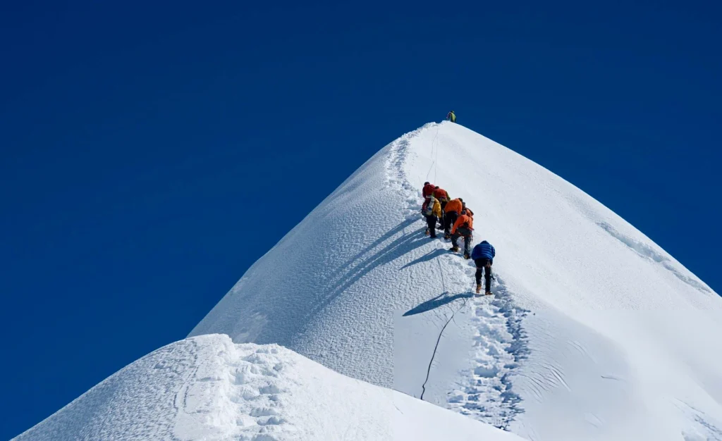 A line of mountaineers in colorful winter gear trekking up a steep, snowy mountain ridge toward the summit under a clear blue sky.