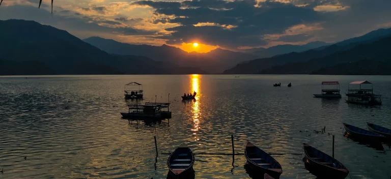 Golden sunset over Phewa Lake in Pokhara, Nepal, with traditional wooden boats floating on the calm water against a backdrop of silhouetted mountains.