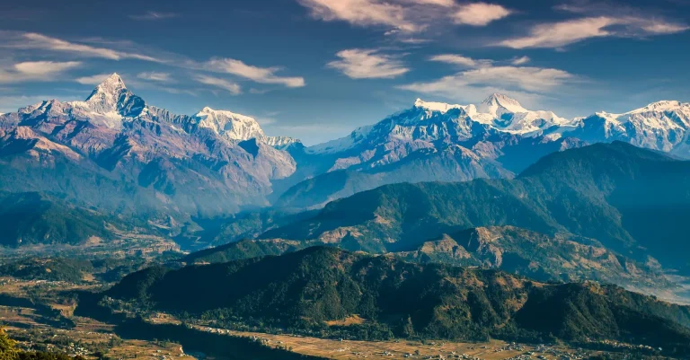 Panoramic view of the Annapurna mountain range and Machhapuchhre (Fishtail peak) over the Pokhara valley hills in Nepal under a partly cloudy blue sky.