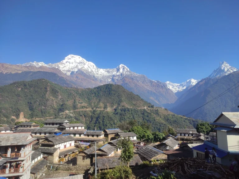 A stone-roofed village in Ghandruk, Nepal, set against the snow-capped peaks of Annapurna South and Machhapuchhre (Fishtail Mountain) under a clear blue sky.