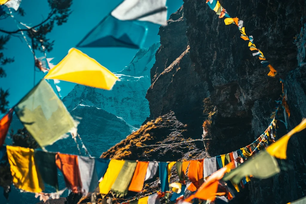 Colorful Tibetan prayer flags flutter in the foreground, framing a steep rocky mountain face and a distant snow-capped peak under a clear blue sky.