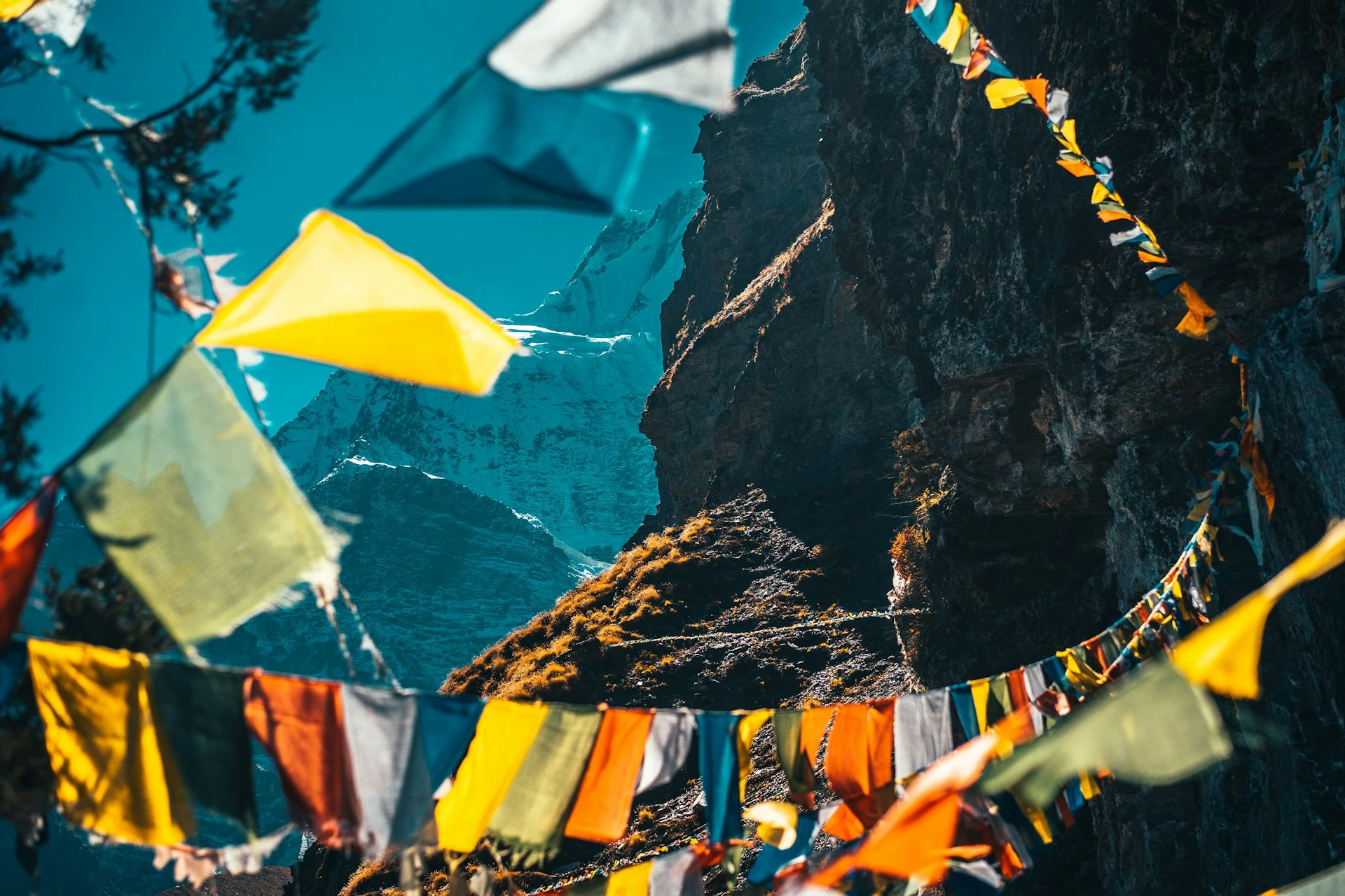 Colorful Tibetan prayer flags flutter in the foreground, framing a steep rocky mountain face and a distant snow-capped peak under a clear blue sky.