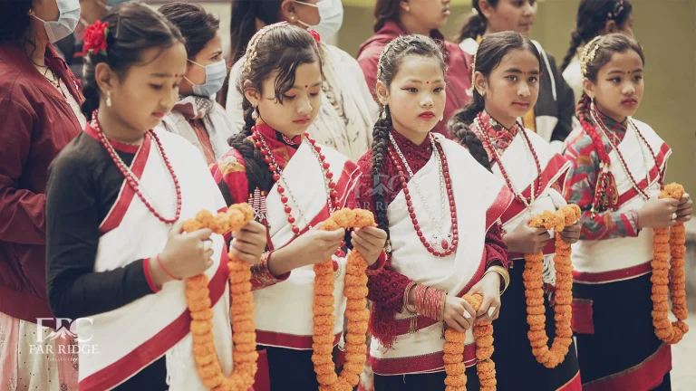 Five young girls representing Panchakanya stand in a row wearing traditional Newar attire (black and red Haku Patasi) while holding marigold garlands for a welcoming ceremony.