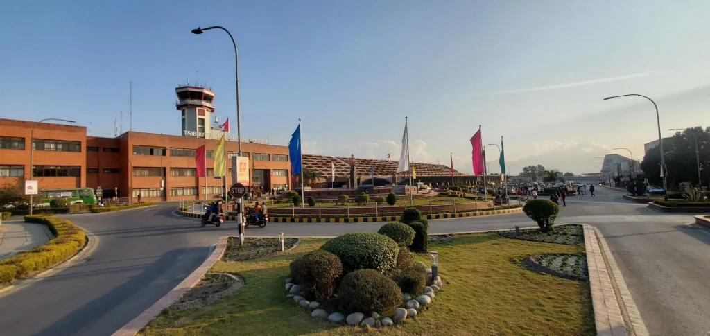 Tribhuvan International Airport terminal building and control tower in Kathmandu, Nepal, surrounded by a roundabout with flags and manicured lawns under a clear sky.